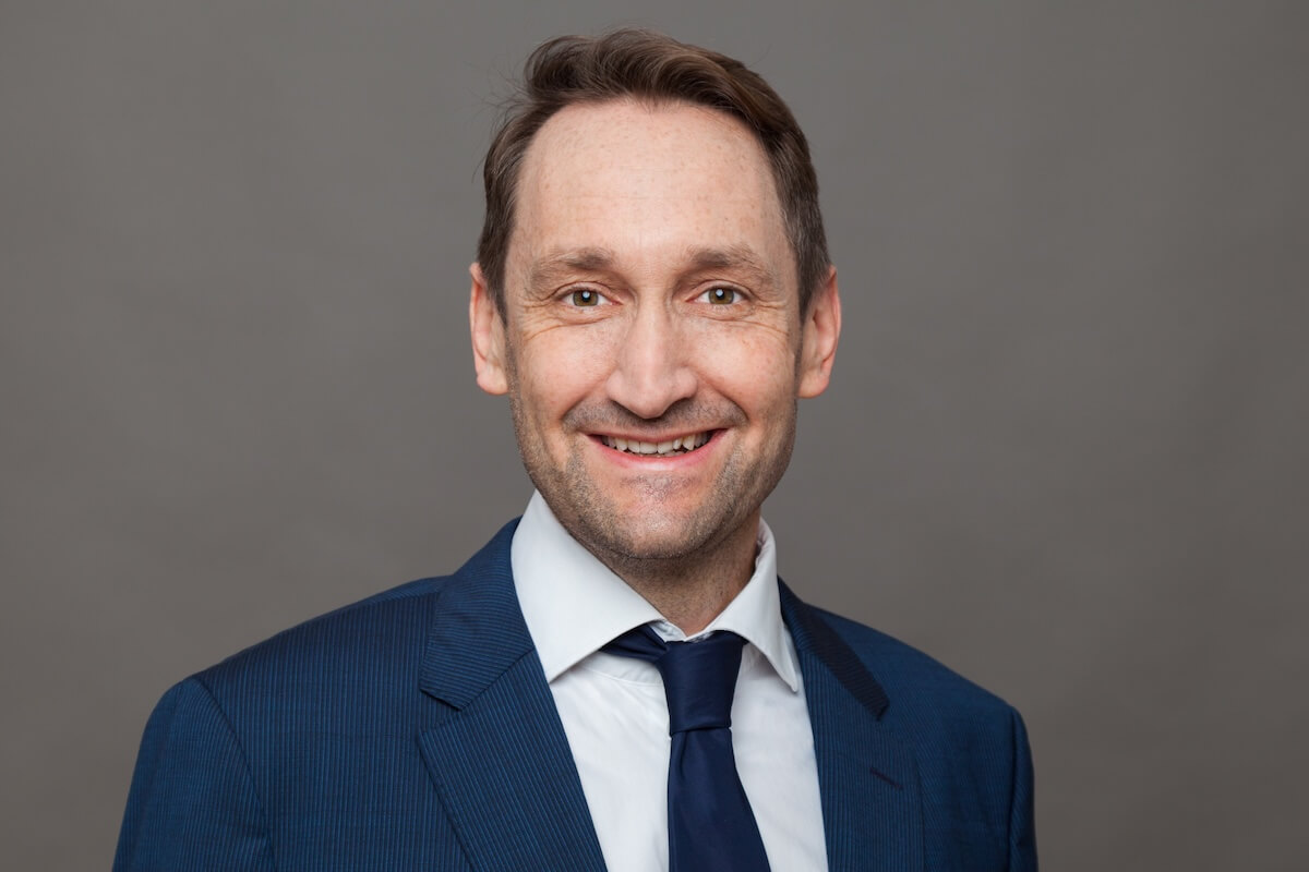 Das Bild zeigt Christian Haase vor einem grauen Hintergrund. Er trägt einen blauen Anzug mit weißem Hemd, dunkelblauer Krawatte und kurzes braunes Haar. 