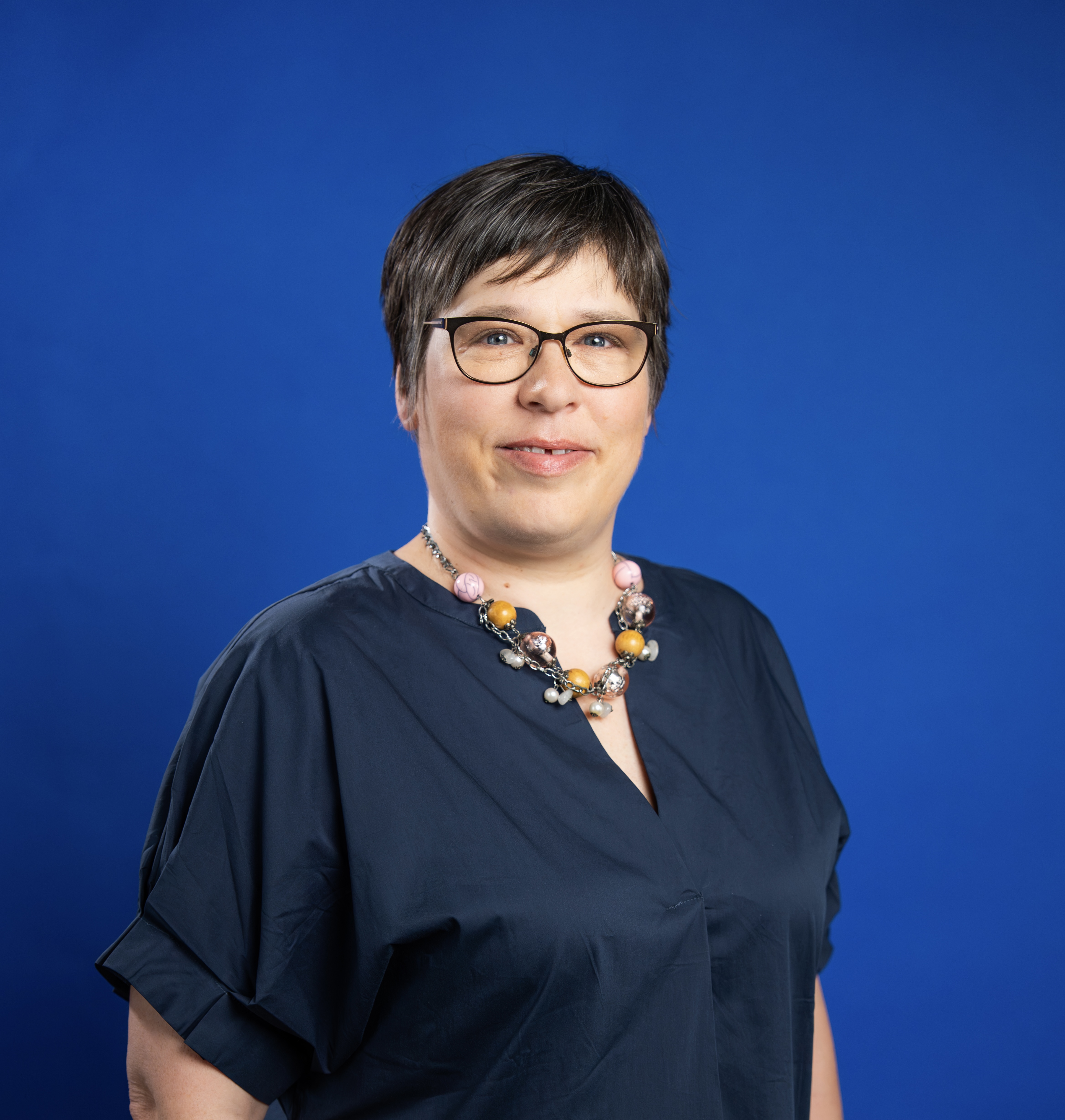 Das Bild zeigt Anja Molnar vor einem dunkelblauen Hintergrund. Sie trägt ein blaues Kleid, eine Brille und hat kurze braune Haare.