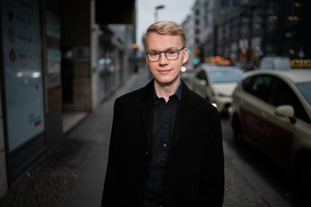 Das Bild zeigt Konrad Klamann vor auf dem Gehweg einer großen Straße. Er trägt ein schwarzes Hemd und ein schwarzes Sakko, sowie eine schwarze Brille und kurze, blonde Haare