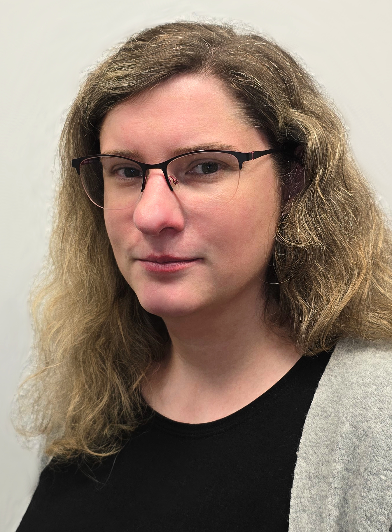 Das Bild zeigt Anne-Katrin Schulze vor einem weißen Hintergrund. Sie trägt langes, blondgelocktes Haar, ein schwarzes Shirt, eine graue Jacke und eine schwarze Brille