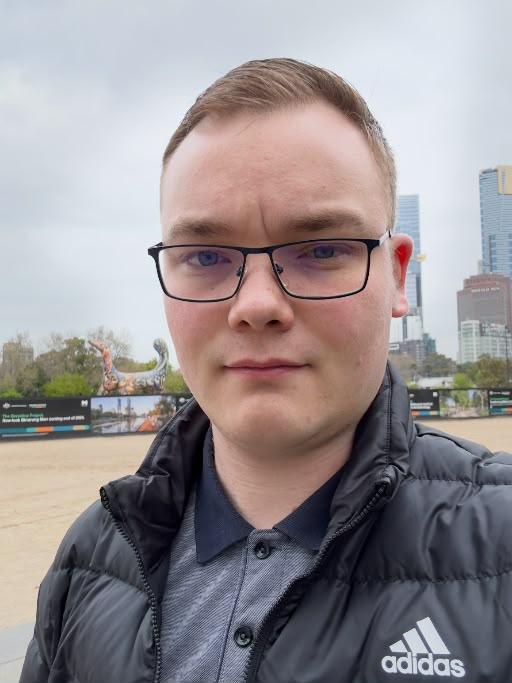 Das Bild zeigt Dominik Sussmann auf einem großen Platz. Er trägt ein blaues Shirt, eine schwarze Weste, kurzes braunes Haar und eine schwarze Brille.