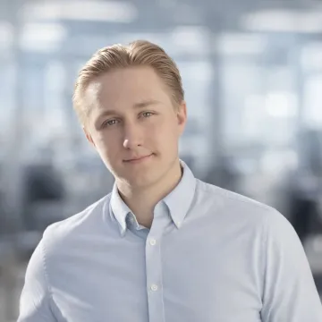 Das Bild zeigt Benjamin Dahler vor einem bewegten Hintergrund. Er trägt ein hellblaues Hemd und kurze blonde Haare.