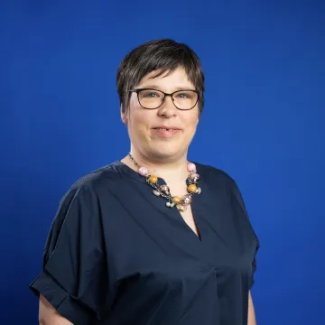 Das Bild zeigt Anja Molnar vor einem dunkelblauen Hintergrund. Sie trägt ein blaues Kleid, eine Brille und hat kurze braune Haare.
