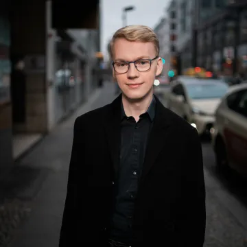 Das Bild zeigt Konrad Klamann vor auf dem Gehweg einer großen Straße. Er trägt ein schwarzes Hemd und ein schwarzes Sakko, sowie eine schwarze Brille und kurze, blonde Haare