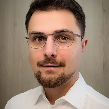 Das Bild zeigt Rune Betz. Er trägt ein weißes Hemd, Drei-Tage-Bart, eine schwarze Brille und kurzes, dunkelbraunes Haar