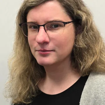 Das Bild zeigt Anne-Katrin Schulze vor einem weißen Hintergrund. Sie trägt langes, blondgelocktes Haar, ein schwarzes Shirt, eine graue Jacke und eine schwarze Brille