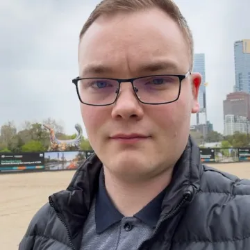 Das Bild zeigt Dominik Sussmann auf einem großen Platz. Er trägt ein blaues Shirt, eine schwarze Weste, kurzes braunes Haar und eine schwarze Brille.